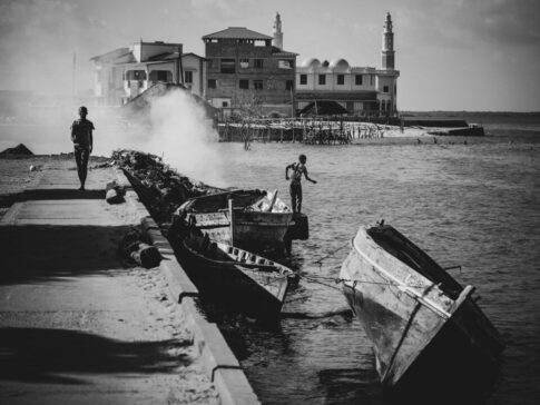 A black and white image of a coastal scene with three wooden boats by a pier, a person walking, smoke in the background, and buildings, including a mosque with domes, across the water.